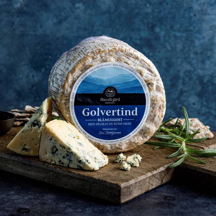 Et stykke blåmuggost med urter ved siden av på en skjærefjøl, med en ostekasse i bakgrunnen.