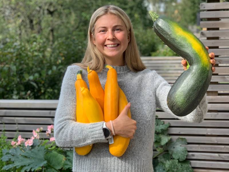 En person holder flere gule squash og en stor grønn zucchini utendørs med blomster og trær i bakgrunnen.