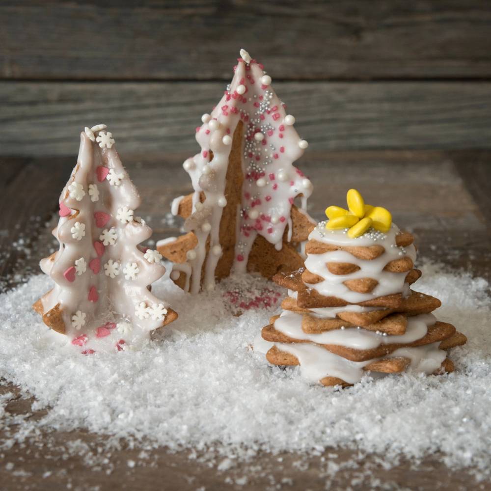 Barn har pyntet pepperkaker til jul med hvit glasur og rosa og hvite strøssel, og det ligger kunstig snø på bordet.
