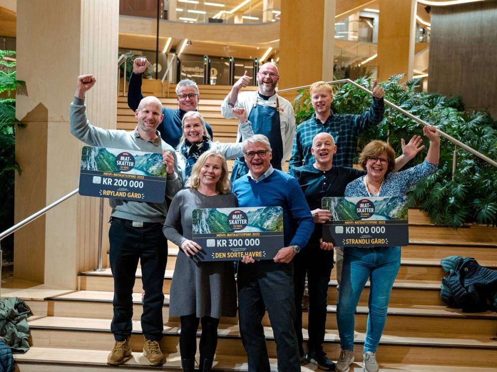 En gruppe mennesker står på en trapp i et innendørs område og holder store sjekk- eller plakater med beløp, med noen som hever armene i triumf.