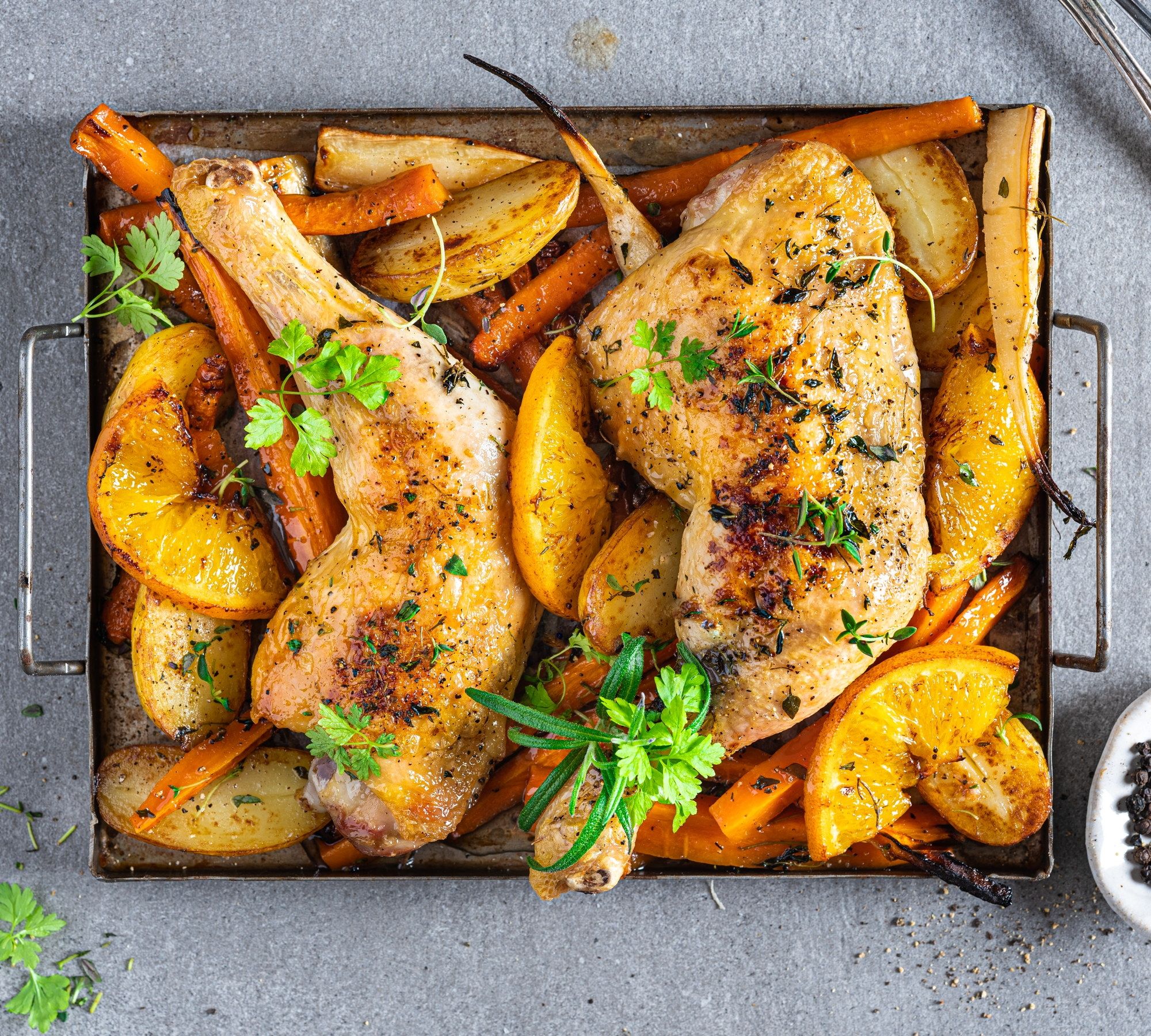 Ovnsbakte kyllinglår med poteter og gulrøtter, pyntet med frisk persille, på en svart stekeplate.