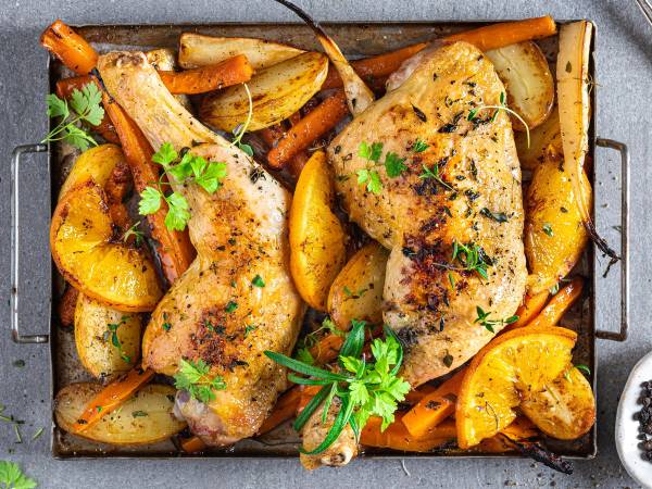 Ovnsbakte kyllinglår med poteter og gulrøtter, pyntet med frisk persille, på en svart stekeplate.