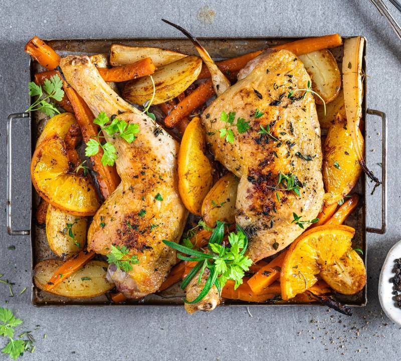 Ovnsbakte kyllinglår med poteter og gulrøtter, pyntet med frisk persille, på en svart stekeplate.