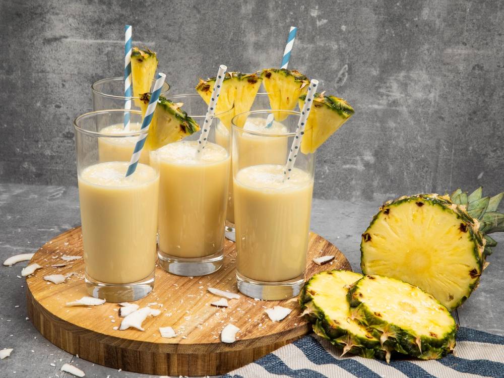 Tre glass med ananasdrikker pyntet med ananasbiter og sugerør, plassert på et rundt trestykke med noen ananasbiter ved siden av, mot en grå bakgrunn.