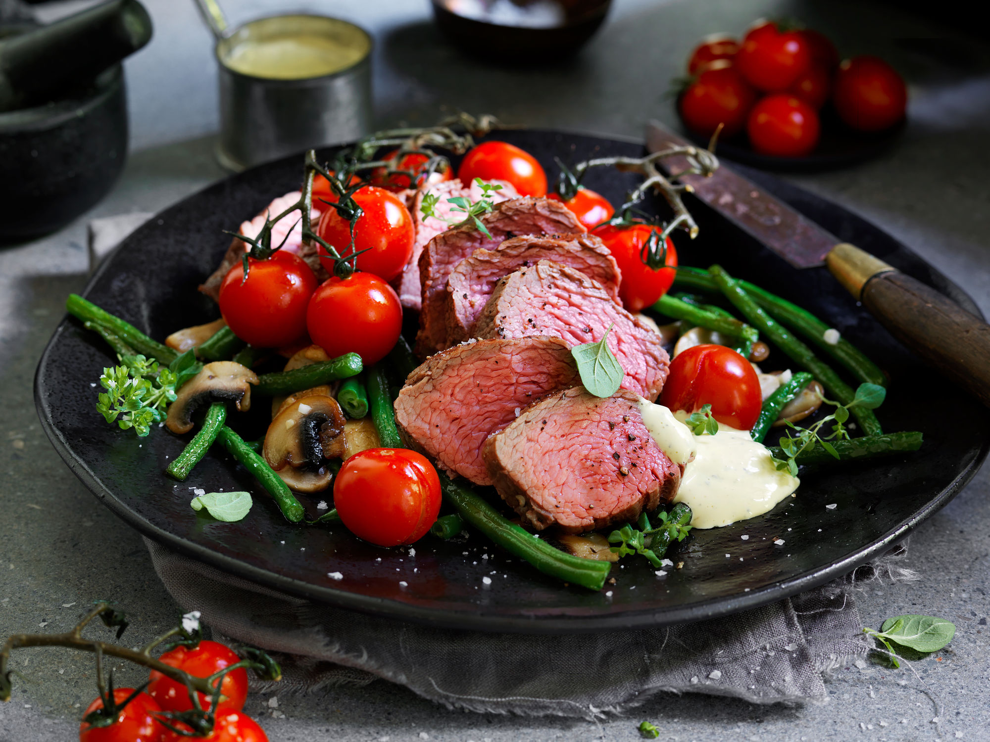 Biff med tilbehør som grønne bønner, cherrytomater og sopp på en svart tallerken.