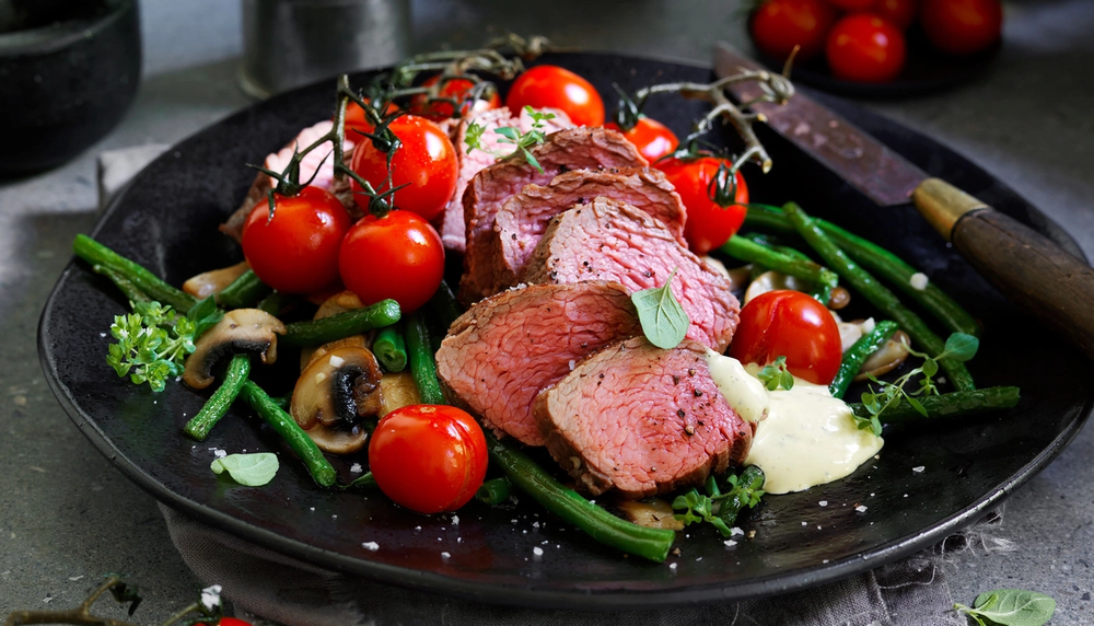 Biff med tilbehør som grønne bønner, cherrytomater og sopp på en svart tallerken.