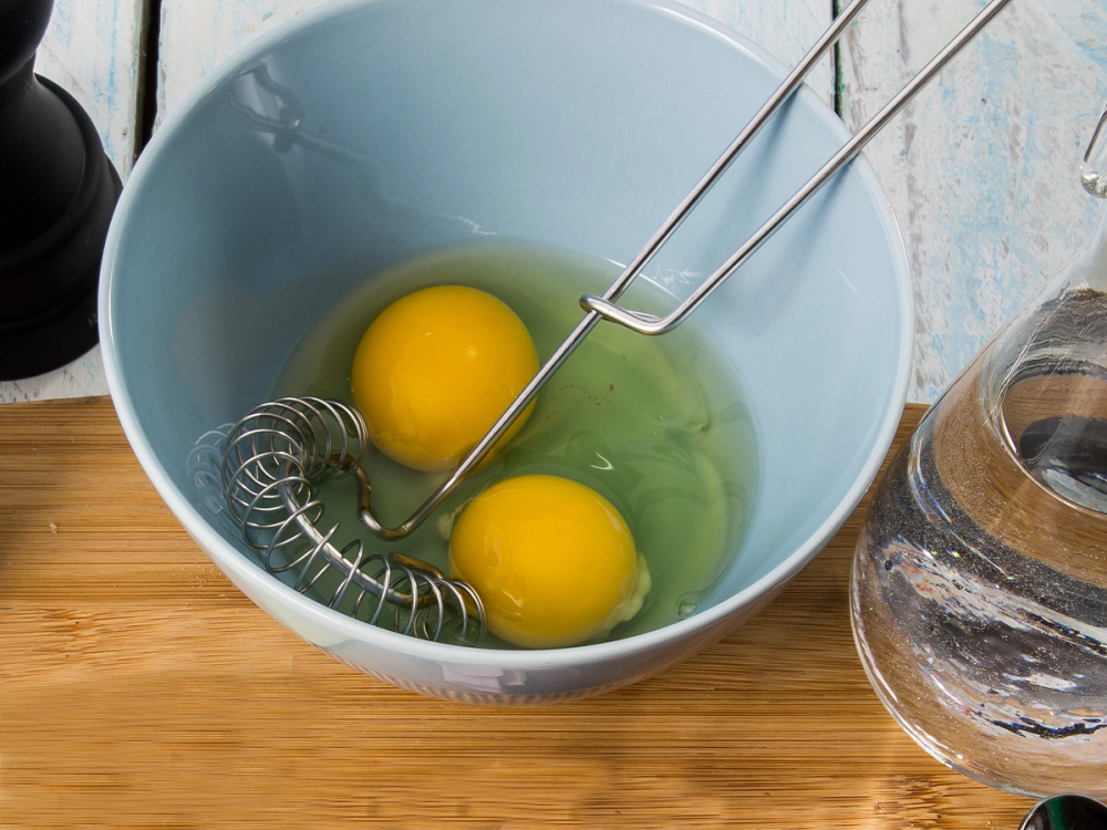 To egg i en blå bolle med en visp og en metallkrok, klar til å lage omelett.