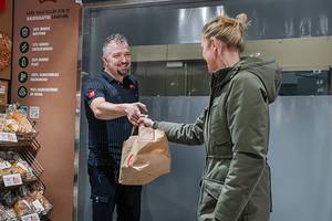 En person i grønn jakke mottar en matpose fra en ansatt i en butikk eller kafé, med en meny eller informasjonstavle i bakgrunnen.