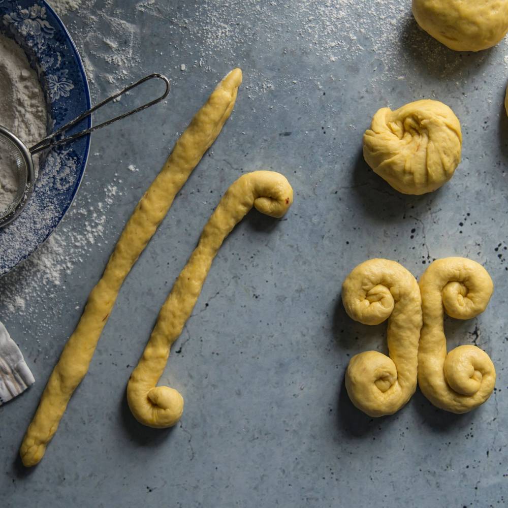 Lussekattformer formet som kvadrater og spiraler på et kjøkkenbord før baking.