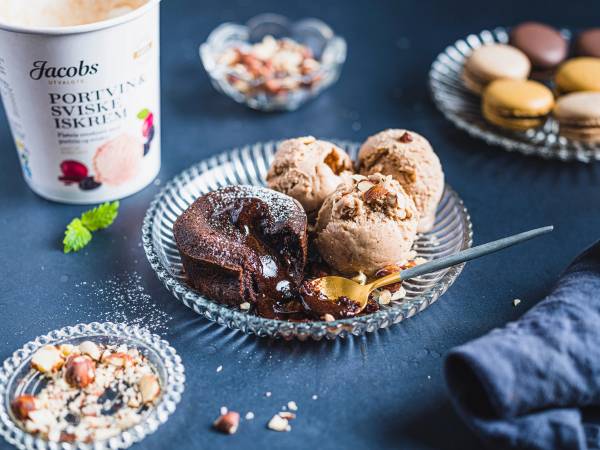 Ulike iskuler på et glassfat med en skje, omgitt av små kaker og en kopp med Jacobs iskrem, på en blå bordduk.