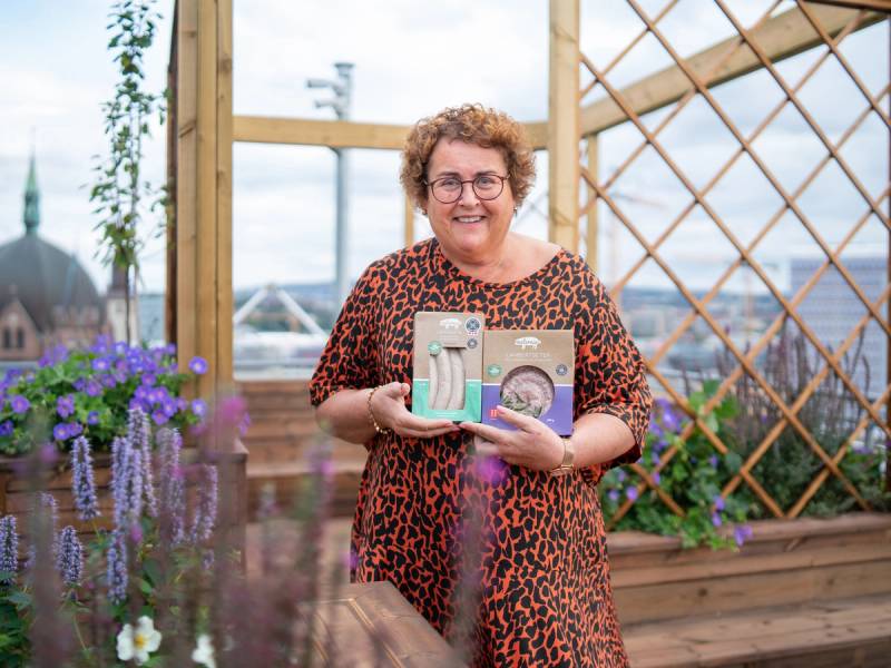En kvinne holder opp to priser eller utmerkelser på en utendørs terrasse med blomster og treverk, med by og kirketårn i bakgrunnen.