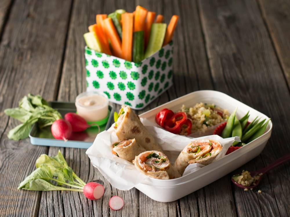 Lompe med røkelaks, grønnsaker og dipp i en matboks på et bord med grønnsaker og en kopp med grønnsaker i bakgrunnen.