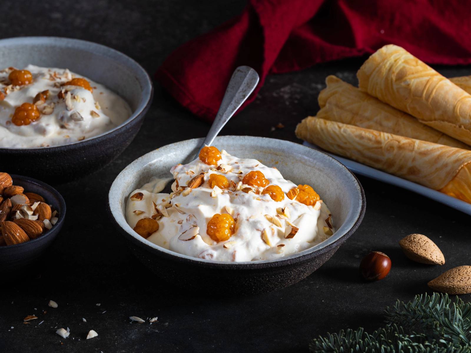 Multekrem i en skål med sprøytetopping og nøtter, omgitt av vafler og julekuler på et mørkt bord.