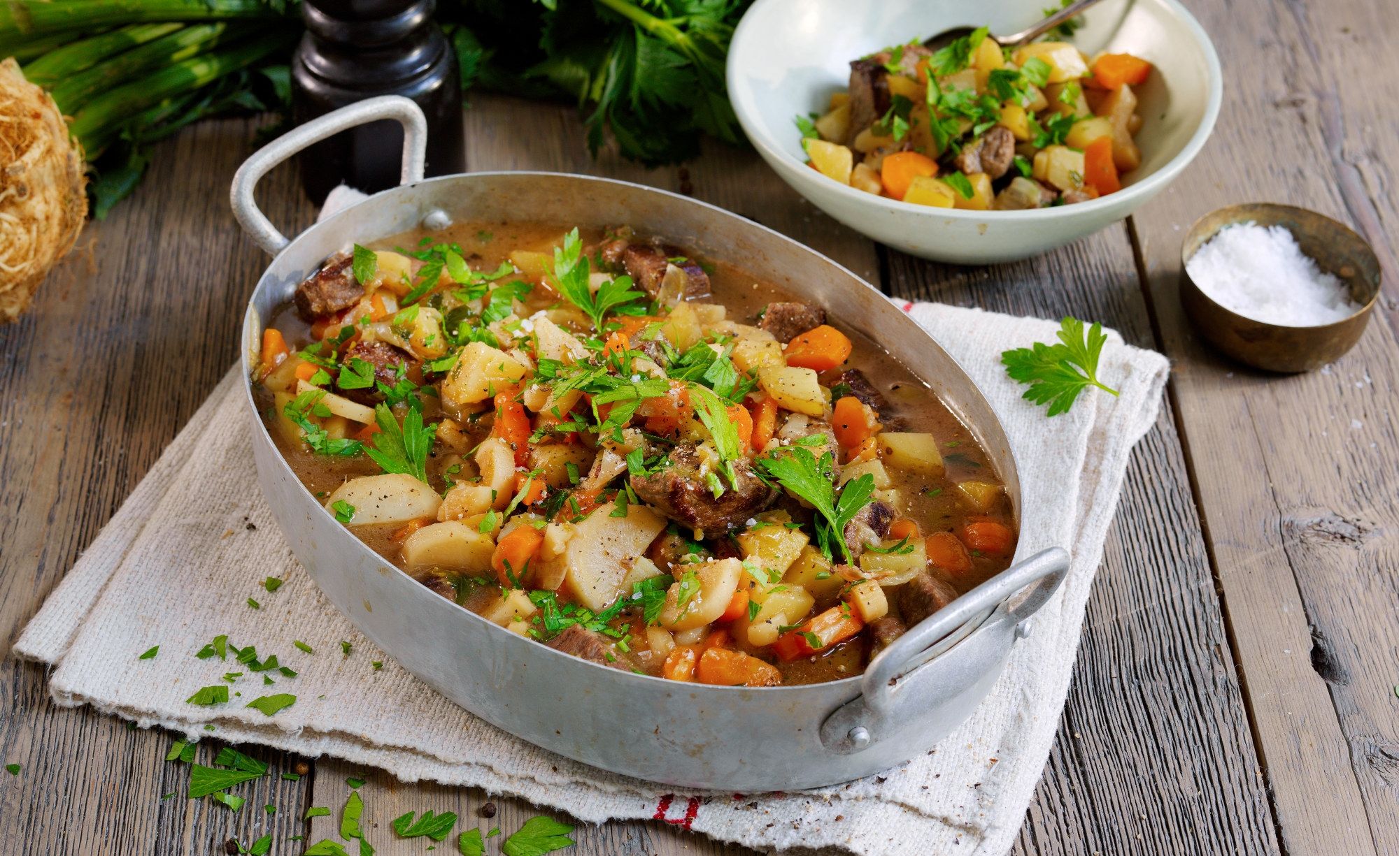Lapskaus i en stor gryte med grønnsaker og kjøtt, servert med en liten bolle salt og en skje med salt ved siden av.