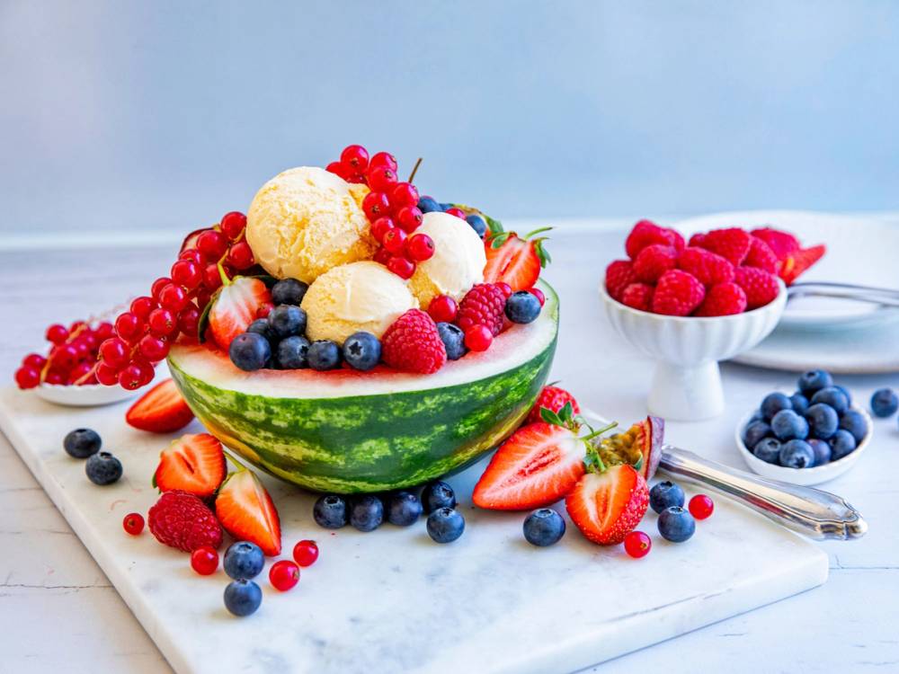 Vannmelonkake dekorert med vaniljeis og ferske bær, omgitt av ekstra bær på et hvitt fat og bord.