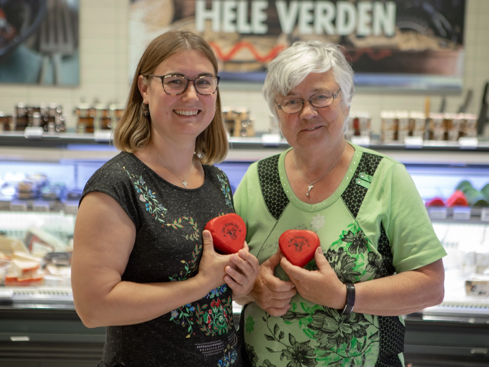 Mor og datter står sammen i en ostebutikk og holder hver sin ost, begge smiler.