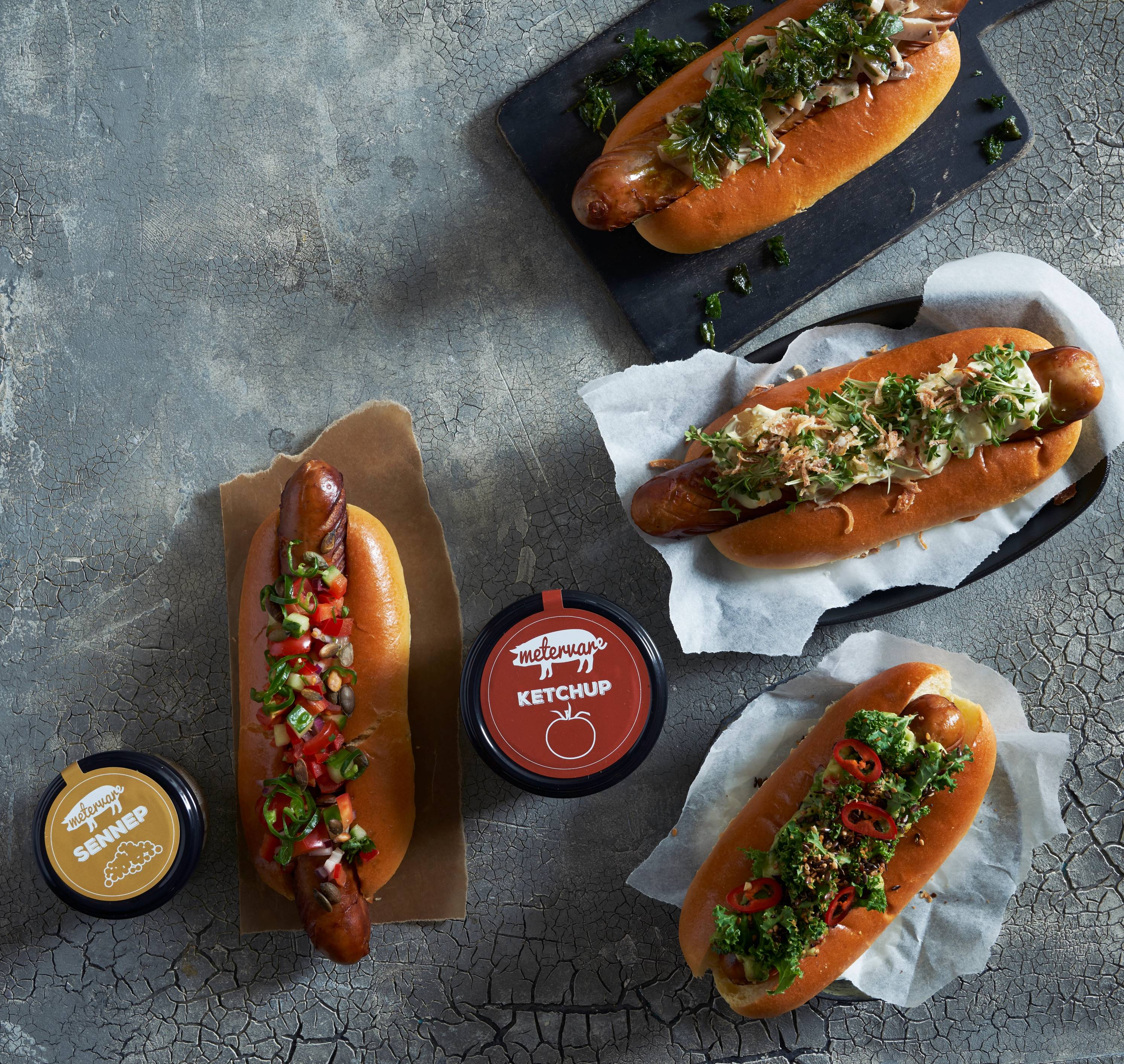 Fire pølser i brød med ulike toppings og en kopp med ketchup på et bord.