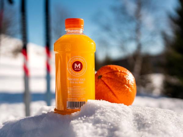 En flaske med oransje drikke og et brunt brød ligger på snø utendørs med en bakke av snø og en blå himmel i bakgrunnen.