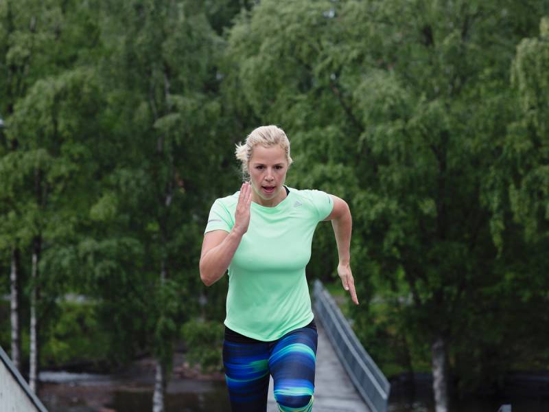 En person løper utendørs på en bro med grønt løv i bakgrunnen, iført lys grønn t-skjorte og blå tights.