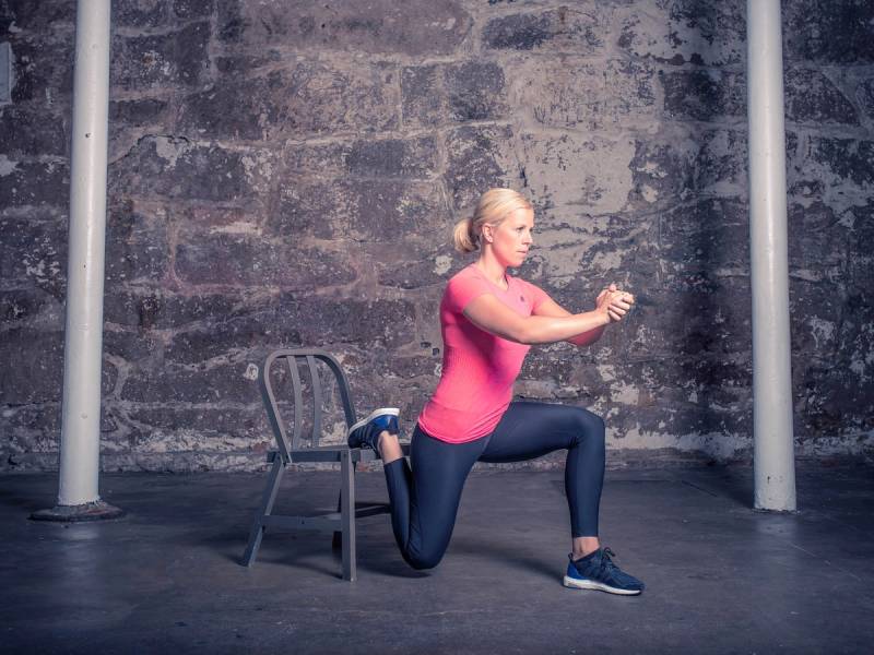 En person utfører en splittknebøy med ett kne på en stol foran en grå murvegg, iført rosa topp og mørke bukser.