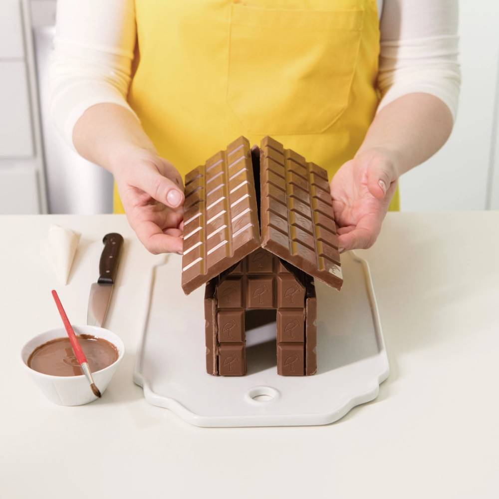 Person som holder et byggesjokoladehus med tak laget av sjokoladeplater på en hvit skjærefjøl, med en bolle med smeltet sjokolade og en rød strikkepinne ved siden av.