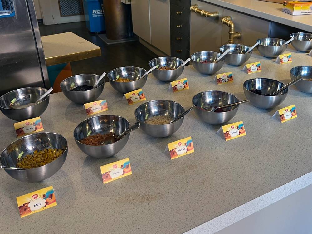 Ti metallskåler med ulike typer frø eller korn, plassert på en benkeplate med små etiketter foran hver skål, i et arbeidsmiljø.