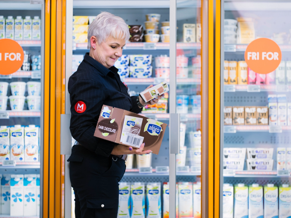 En ansatt i butikk som holder flere matvarer og står foran en kjøledisk med melk og meieriprodukter, med et skilt som sier 'Fri for'.
