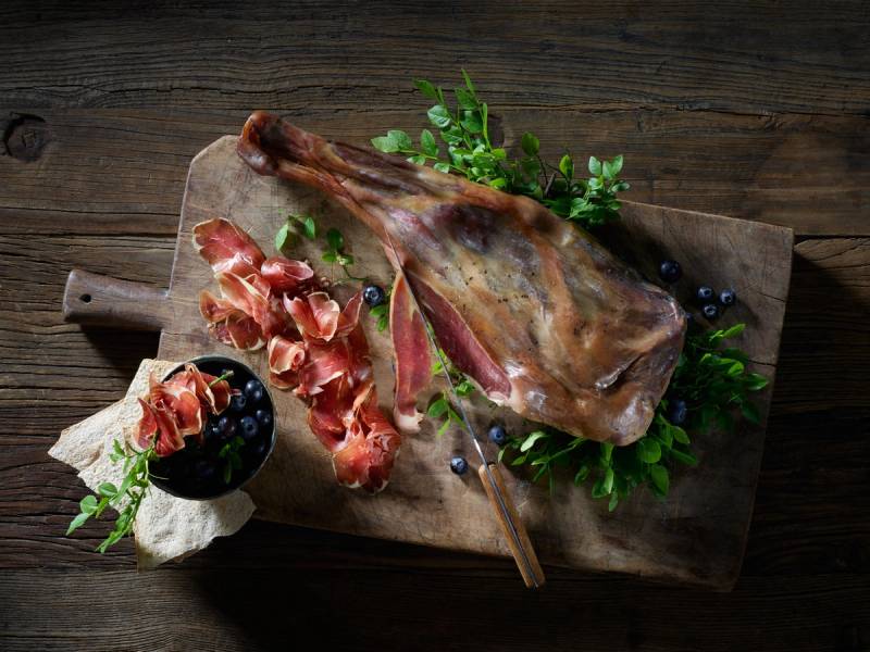 Speket lammekotelett med skinke og blåbær på en treplate med grønt garnityr, på en mørk trebakgrunn