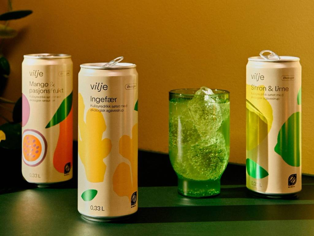 Tre bokser med Vilje-matdrikker og et glass med grønn drikke med isbiter på en bordflate mot en gul bakgrunn.