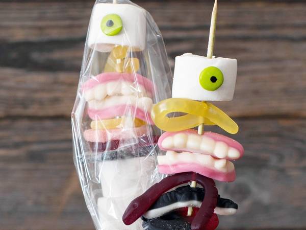 En kreativ halloween-dekorasjon med godteri og ansiktsuttrykk laget av marshmallows og andre søtsaker, plassert i en krukke mot en trebakgrunn.