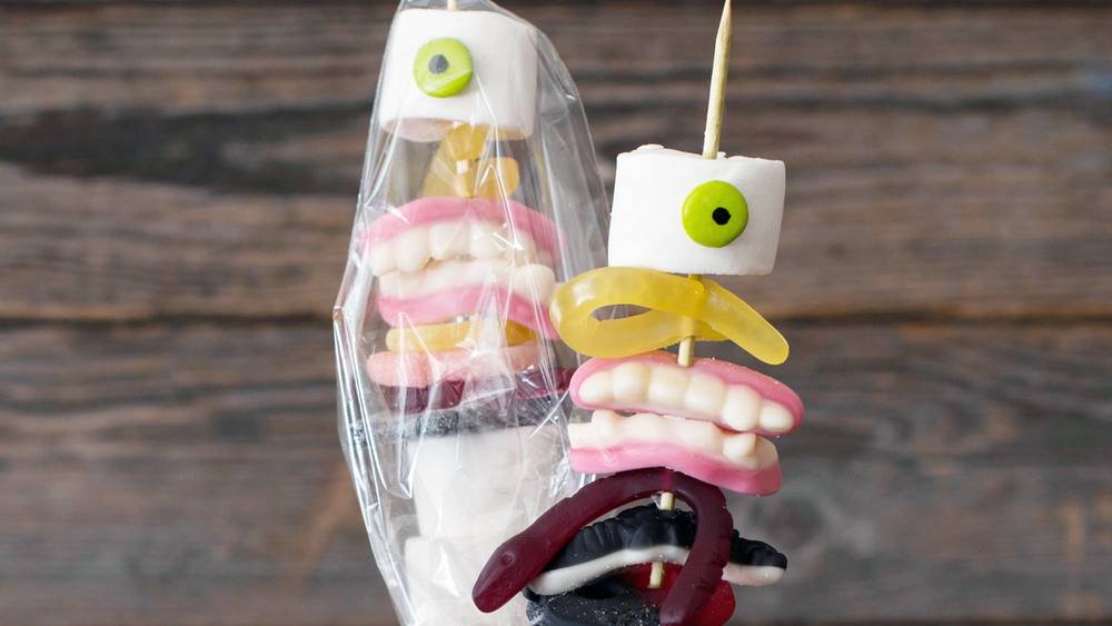 En kreativ halloween-dekorasjon med godteri og ansiktsuttrykk laget av marshmallows og andre søtsaker, plassert i en krukke mot en trebakgrunn.