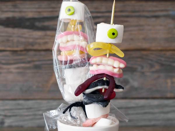 En kreativ halloween-dekorasjon med godteri og ansiktsuttrykk laget av marshmallows og andre søtsaker, plassert i en krukke mot en trebakgrunn.