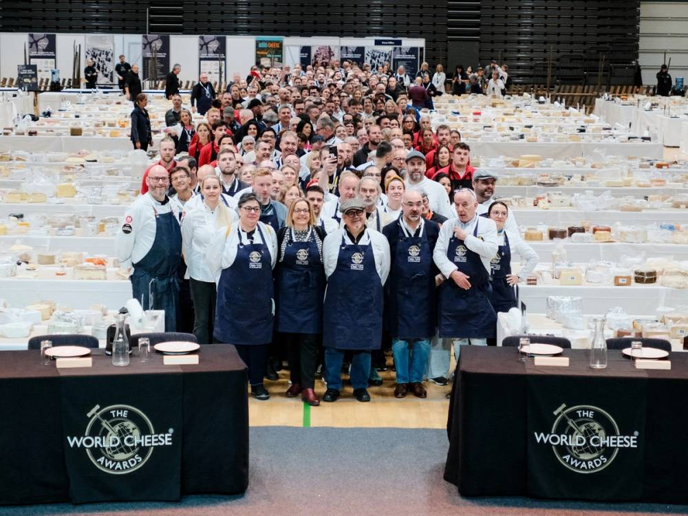 En stor gruppe deltakere på oste-VM står sammen foran langbord med ostekasser i en stor innendørs hall.