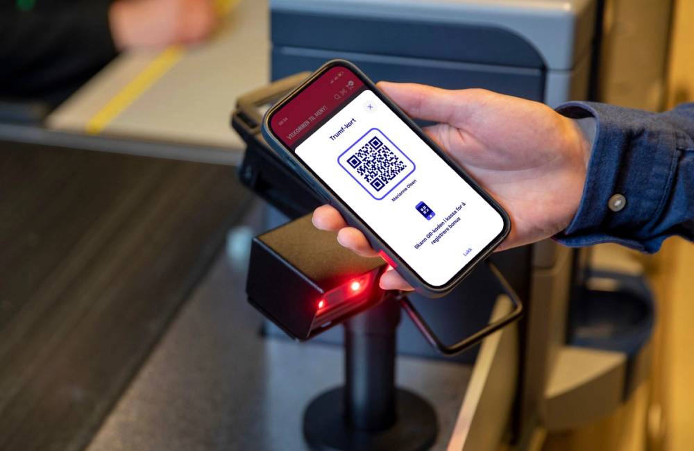 En person holder en smarttelefon med Trumf-kort på skjermen over en leser for kontaktløs betaling, sannsynligvis i en butikk.