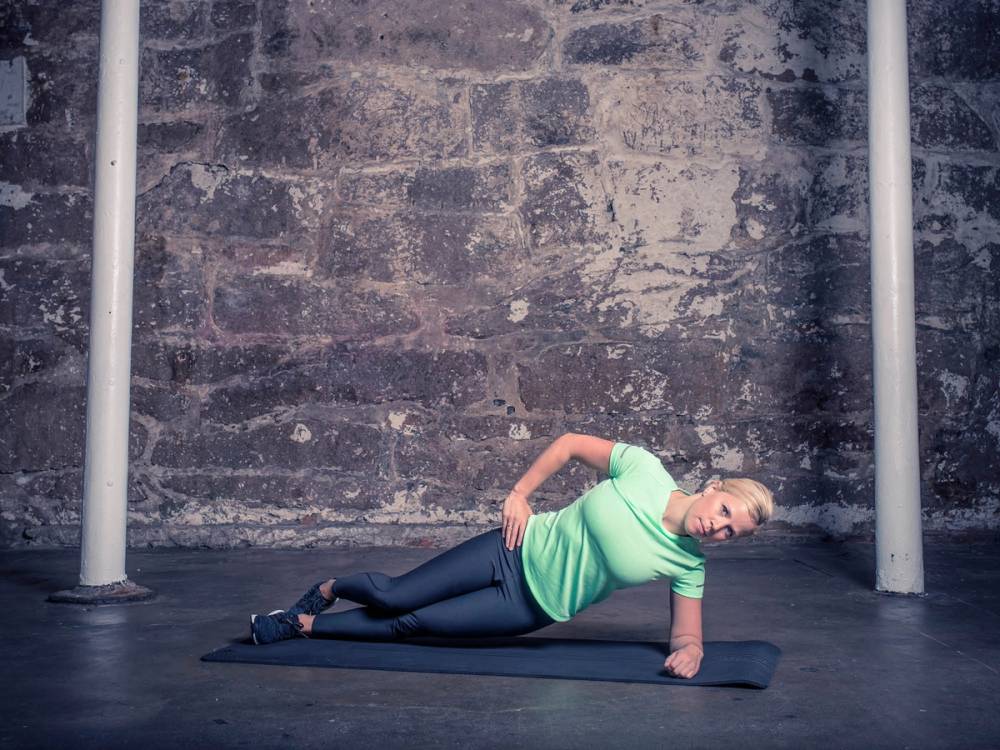 Kvinne gjør en sideplankeøvelse på en matte mot en murvegg, med to stenger på hver side.