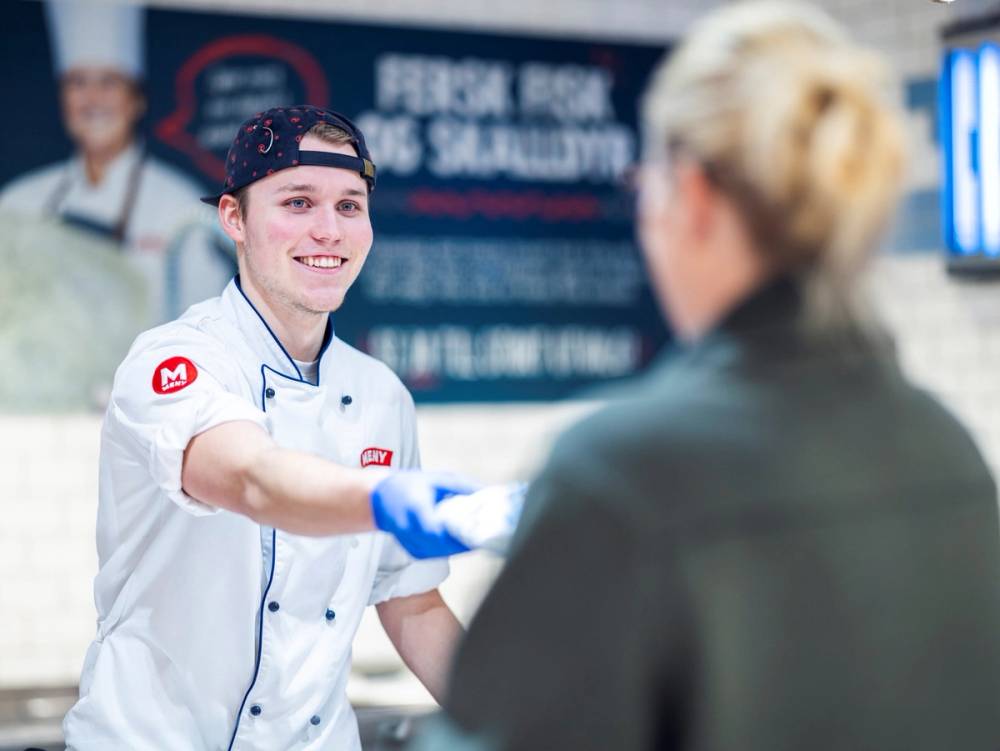 En servitør i hvit uniform med rødt merke gir mat til en kunde på en restaurant eller kafé, med en meny i bakgrunnen.