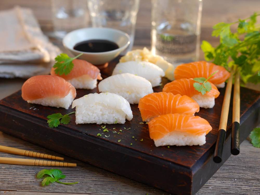 Ulike nigiri-sushibiter med laks og hvit fisk på en svart serveringsplate med soyasaus og grønne urter i bakgrunnen.