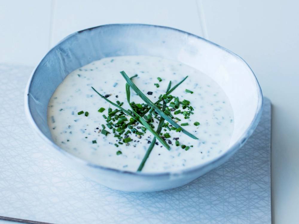 En skål med en hvit saus eller dip pyntet med hakkede grønne urter og to cherrytomater på toppen, plassert på et hvitt underlag.