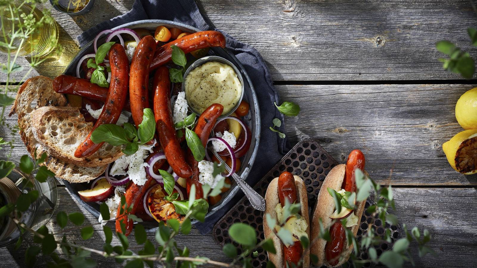 Pølser, brød og tilbehør på et fat, med en skje med saus og grønnsaker rundt, på en trebenk utendørs.