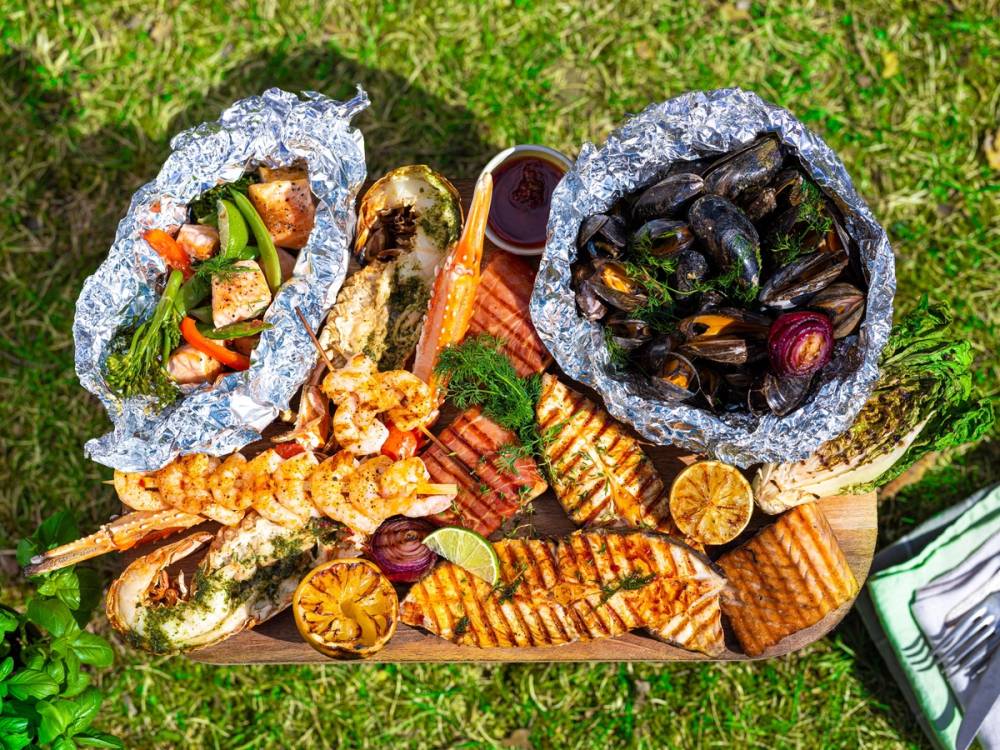 Fisk og skalldyr på et fat med sitronbåter og grønnsaker, delvis innpakket i aluminiumsfolie, på gress.