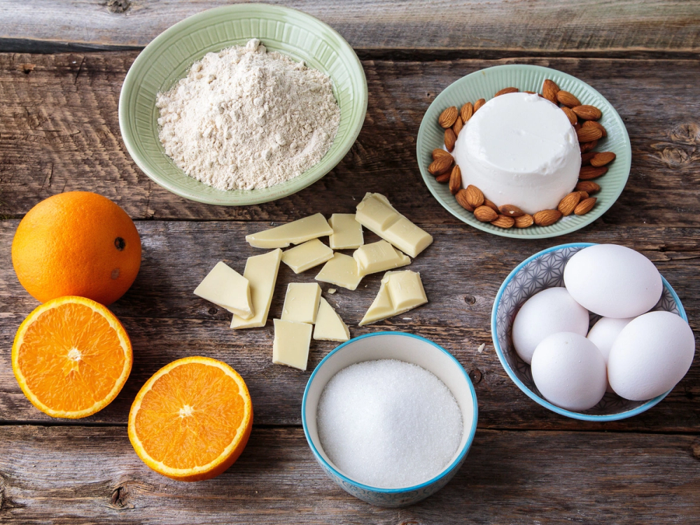 Bilde av ingredienser til baking, inkludert mel, sukker, hvite sjokoladebiter, egg, appelsiner, mandler og kremost, på en treplate.