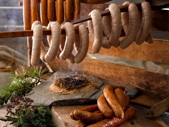 Ulike typer pølser henger til tørk på en trestang, med noen pølser liggende på en stein under, omgitt av grønne planter.