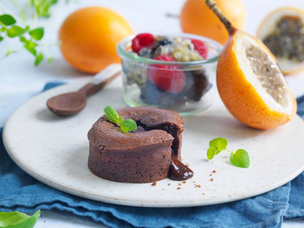 En liten sjokoladekake med granadilla og frukt i en glassbolle, med et halvt granateple og en skje i bakgrunnen.