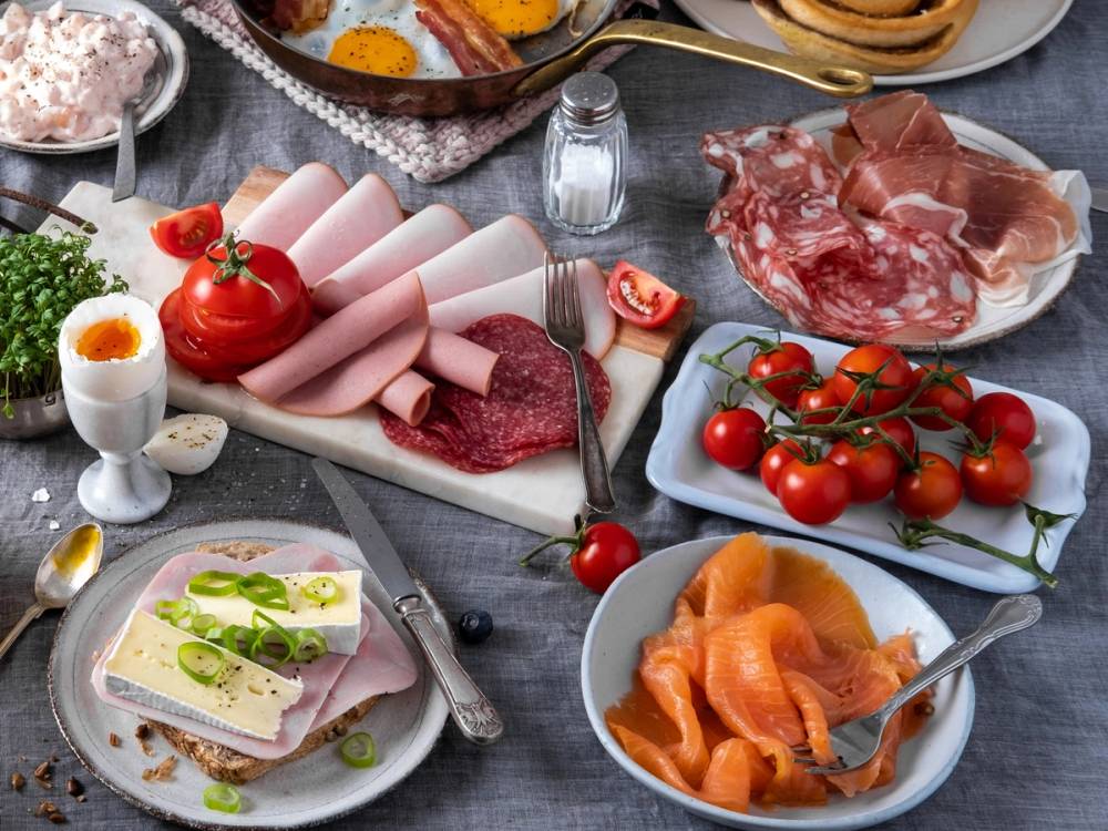 Frokostbord med skinke, salami, tomater, eggerøre, røkelaks, og en skål med smør og grønnsaker.
