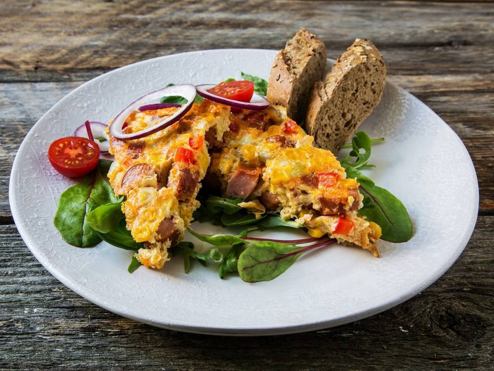 Stekt pølse med ost og grønnsaker på en seng av salat, servert med brød på en hvit tallerken.