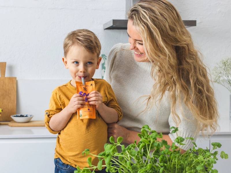En voksen og et barn står sammen i et kjøkken, og barnet holder en pakke med juice eller melk, mens de ser på hverandre.