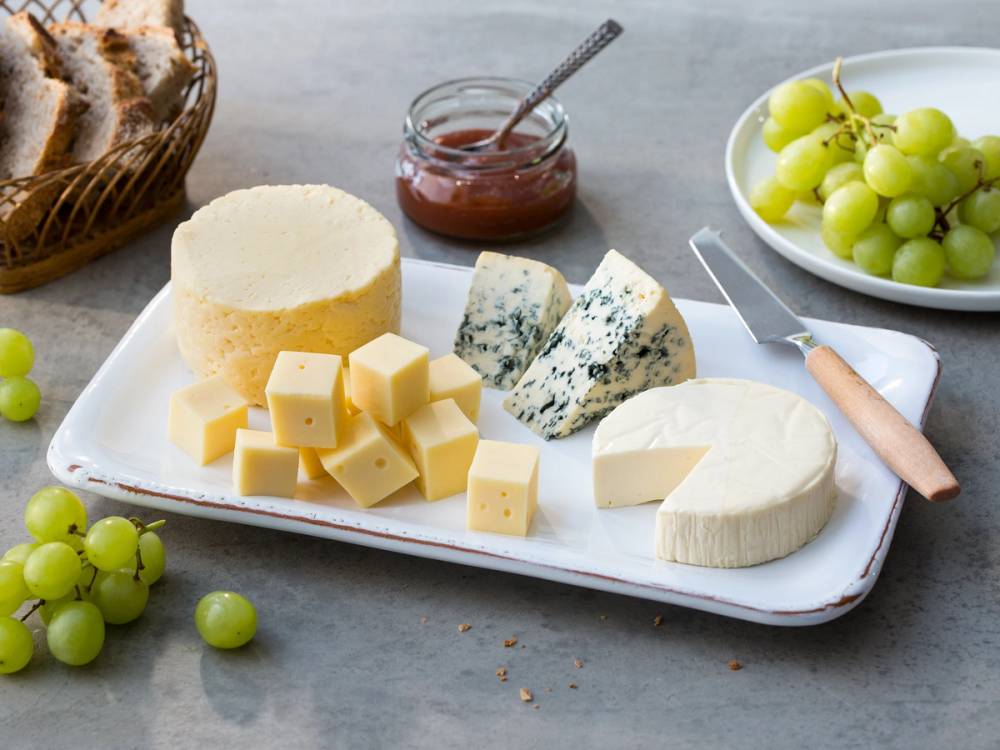 Et ostefat med norske oster, blåmuggost, gulost og hvit ost, omgitt av grønne druer og et glass med rød syltetøy, på en lys bakgrunn.