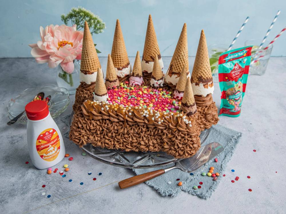 En iskremkake pyntet med flere kjeks og fargerike strøssel, omgitt av en blomsterbukett, en flaske med saus og to stråpinner, på et lyst bord.