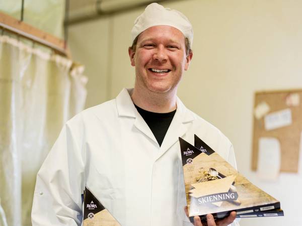 En person i hvit kokkeflate holder to pakker med bakevarer i et rom med beige vegg og oppslagstavle på veggen.