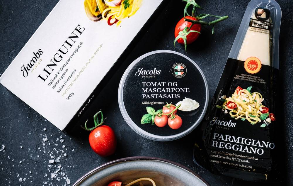 Pakke med Jacobs utvalgte pasta tilbehør, tomat og mascarpone pastasaus, og parmesanreggiano ost på et bord med tomater og krydderurter.